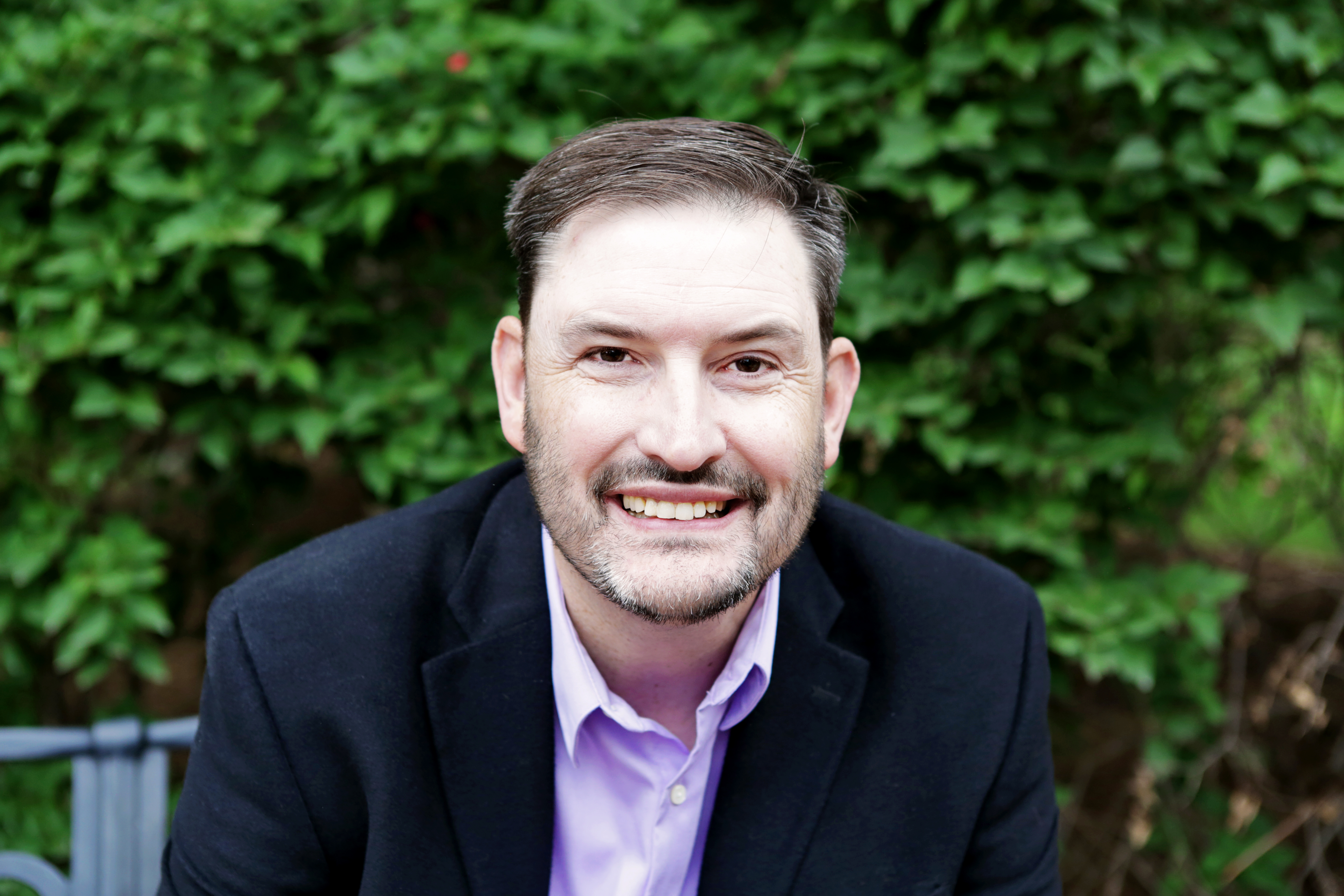 Portrait of Michael Harshbarger, smiling in a dark blazer and light shirt, seated outdoors.